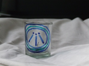 a clear glass candle holder painted with, in blue, the Awen symbol: three line point upwards and inwards to three dots, the whole surrounded by three circles.