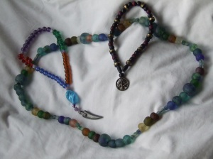 three strings of beads on a cloth. one long one is of recycled bottle glass, uneven beads of green and blue, and the other two are shorter with pendants - a knife blade and a tree.